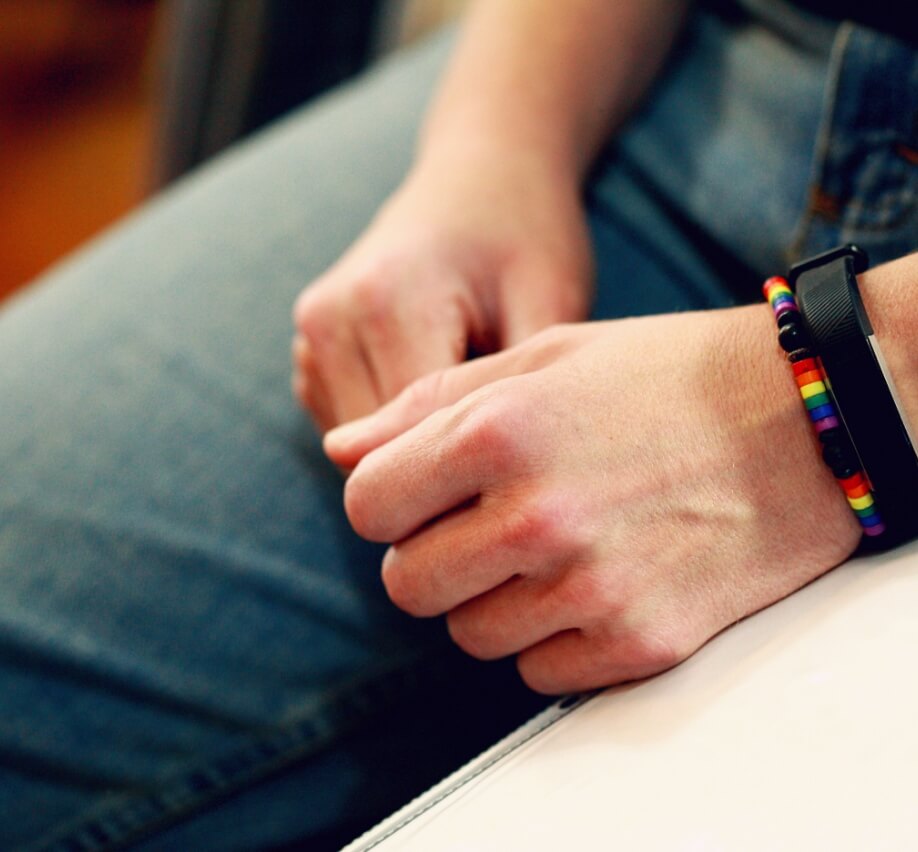 A close-up photo showing a person's clasped hands resting on their lap. One hand has a black smartwatch and a colorful beaded bracelet with a pride flag motif.