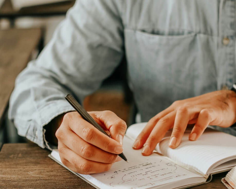 Person in a denim shirt writing notes in a journal with a black pen, focusing intently on the task at hand with a blurred background.