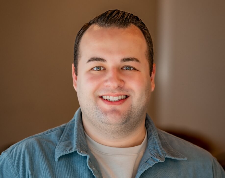 Headshot of Jordan Madden, smiling in a light grey T-shirt and blue denim shirt. The warm, neutral background complements his approachable demeanor.