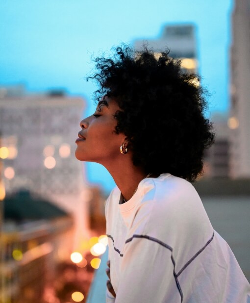 Thoughtful woman with curly hair looking up into the distance, against a blurred cityscape at twilight, evoking a contemplative mood.